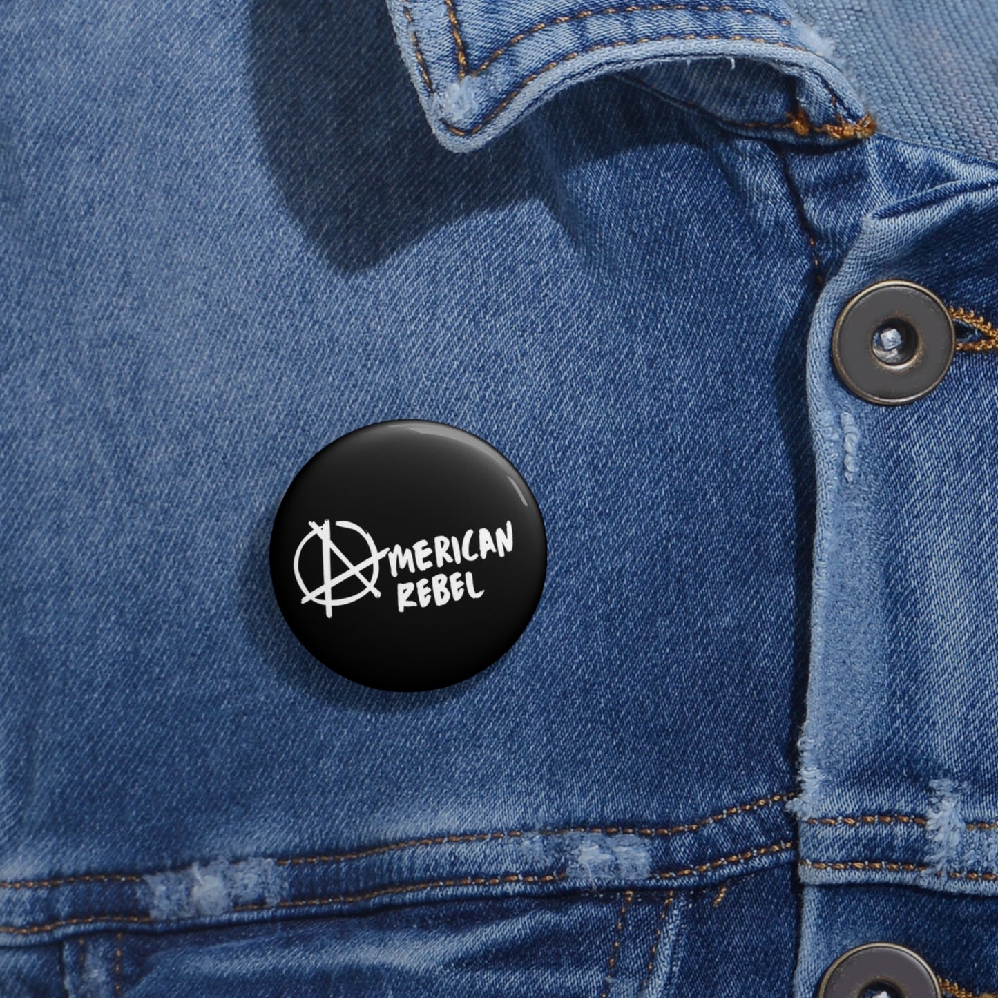 A close-up of a black pin button securely fastened to a blue denim jacket. Proudly featuring an anarchy symbol and the words American Rebel in white, this UV-resistant accessory is assembled in the USA, ensuring lasting defiance against both sun and convention.