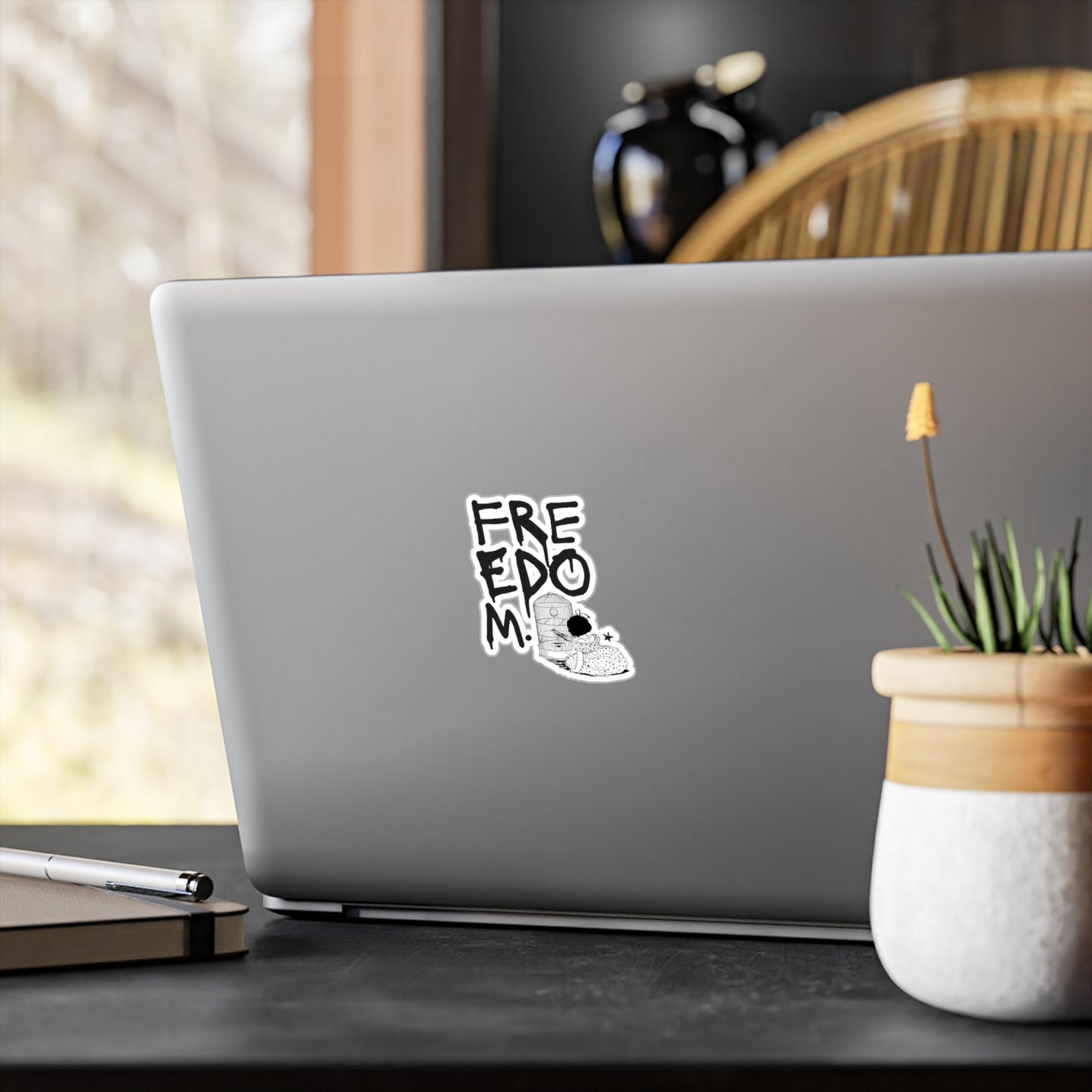 A silver laptop on a dark table near a window with blurred greenery outside features a vinyl decal with FREEDOM and a panda. Nearby, there’s a pen, a plant in a pot, and the chairs wooden backrest partially visible.