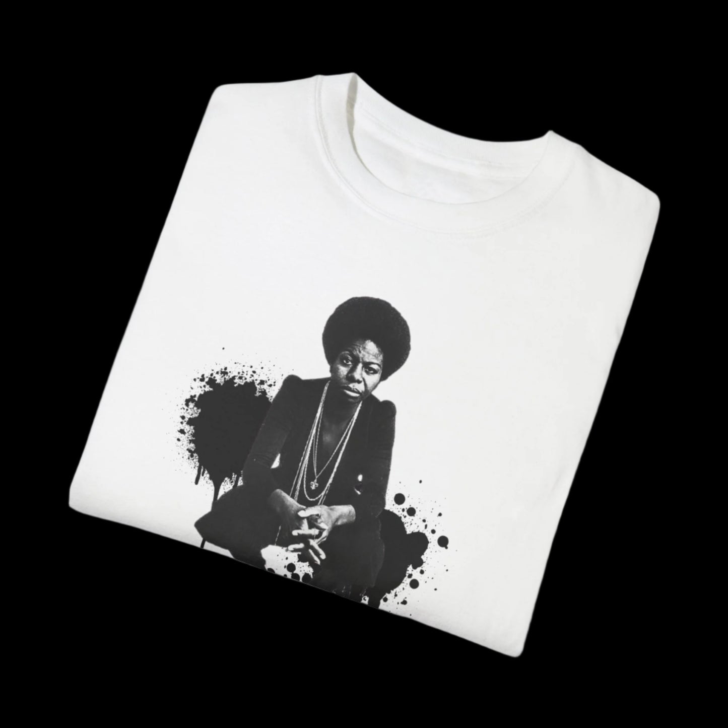A folded white unisex T-shirt with a vintage look features a black and white print of Nina Simone seated, in front of a black paint splatter design, set against a black background.
