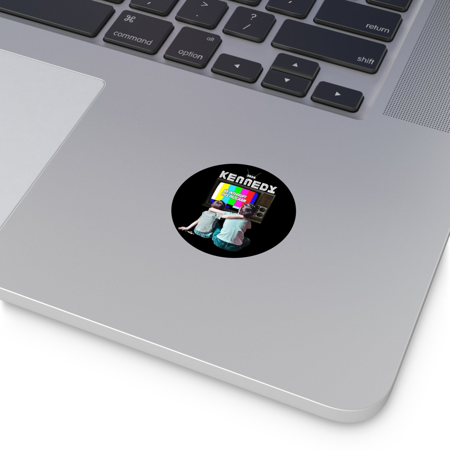 A circular vinyl sticker on a laptop features two kids in front of a retro television set. The word KENNEDY TV is prominently displayed at the top, while the screen beams colorful geometric shapes, and says "We interrupt This Program"