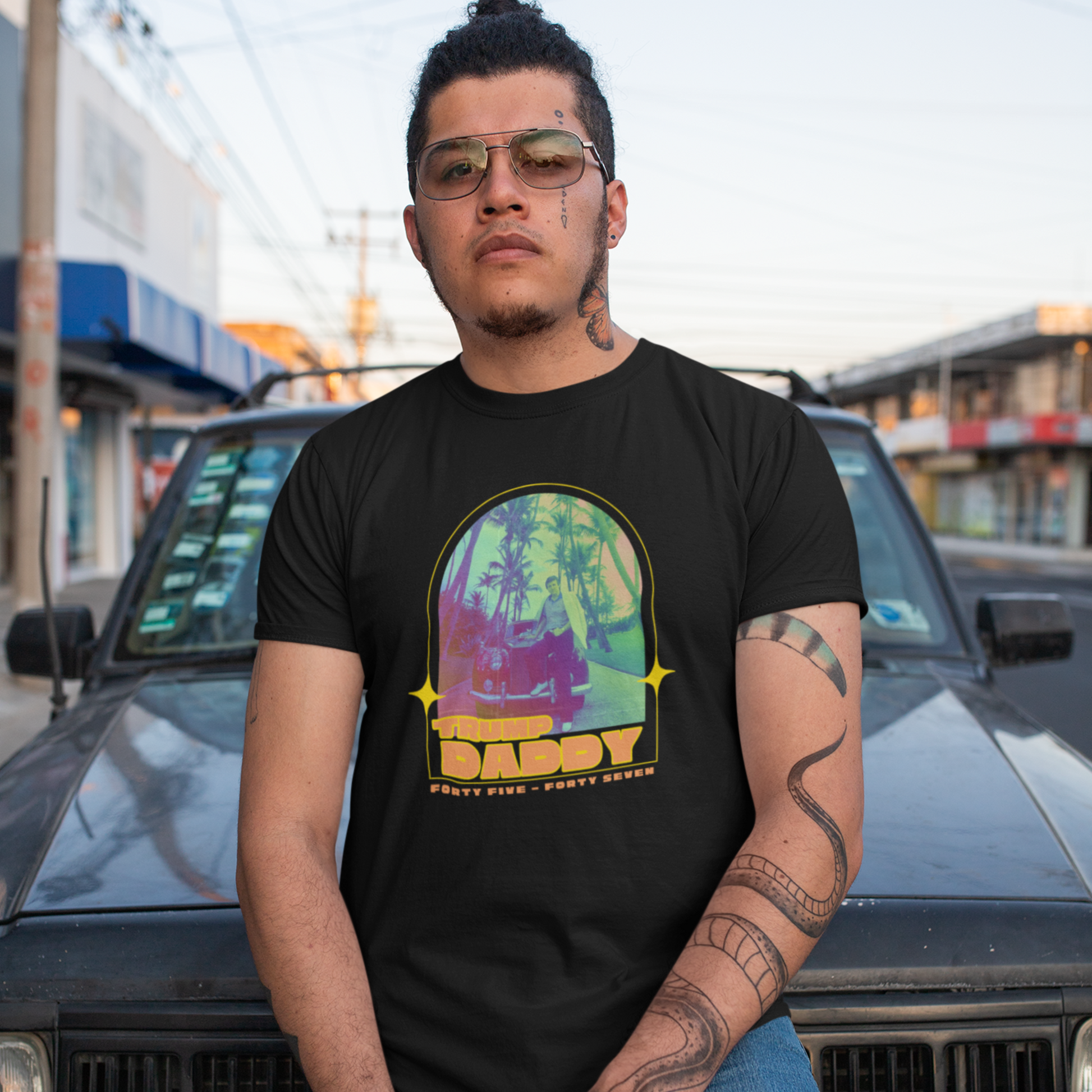 A man, sporting sunglasses and a black retro vintage tee, stands confidently in front of a car on the street. The t-shirt boasts a colorful graphical illustration with the words TRUE DADDY and PARTY EVER written below.
