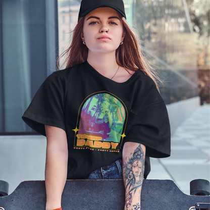 A young woman in a retro vintage tee and black cap holds a skateboard, leaning against a reflective surface. Her shirts colorful graphics burst with vibrancy against the background of lush plants and a stone walkway, encapsulating the timeless appeal of unisex cotton jersey fashion.