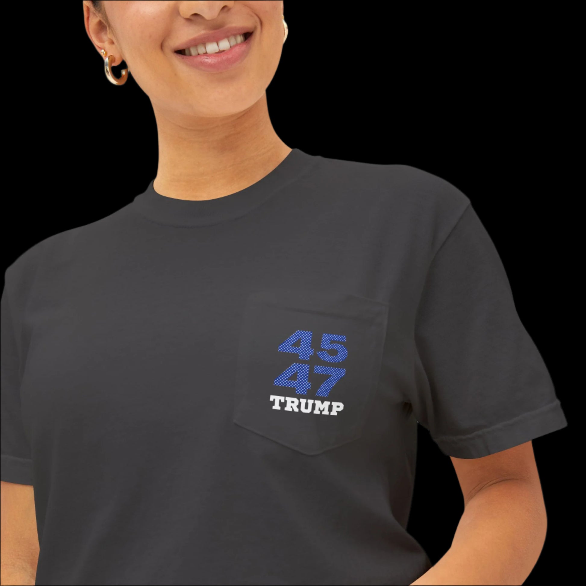 A woman smiling, wearing a garment-dyed black T-shirt. The shirt sports a blue and white design on the pocket area with the numbers 45 47 above the name Trump. The person has gold hoop earrings and hair pulled back