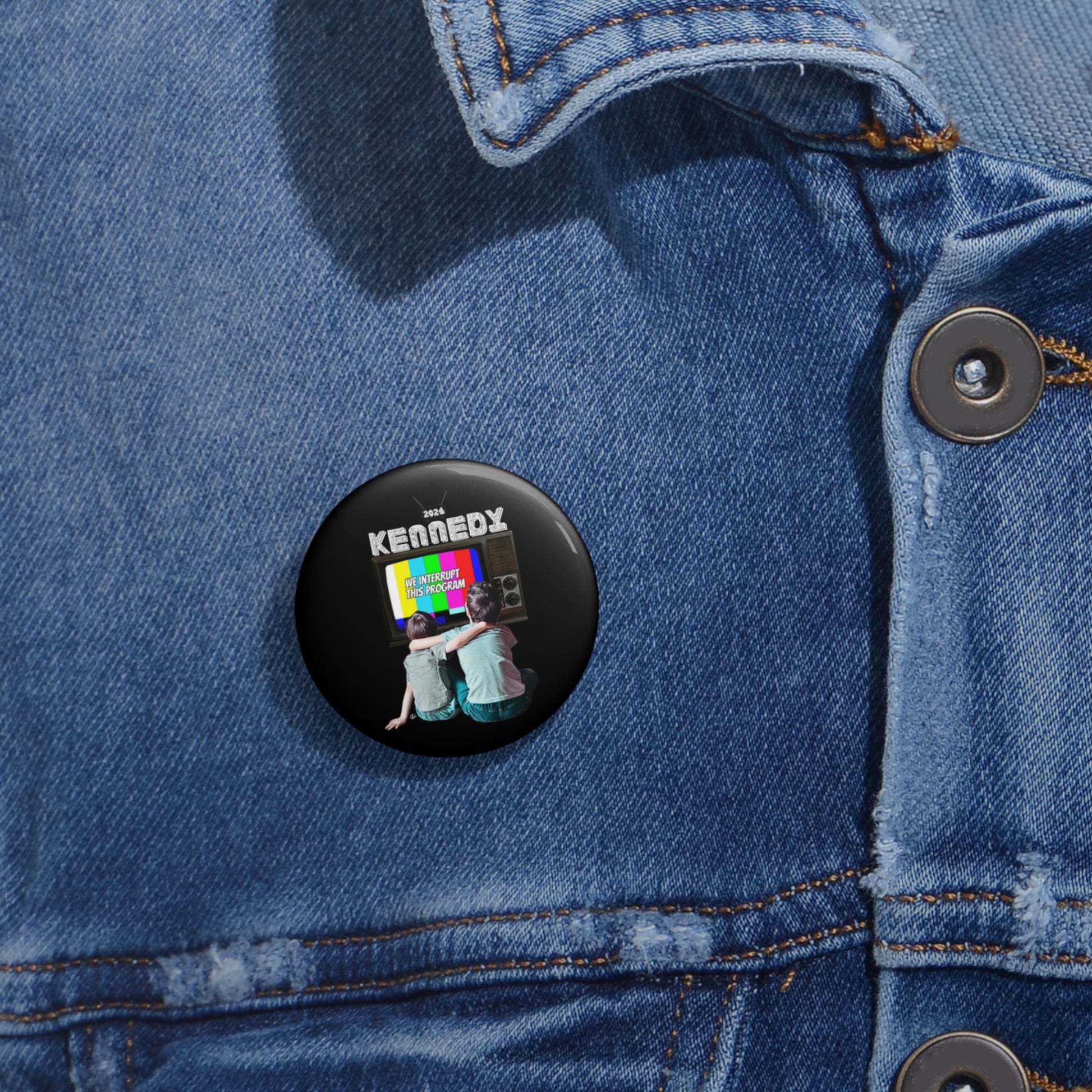 A black button on a denim jacket showcases two people sitting with their backs to a vibrant TV screen, above which KENNEDY is displayed. This RFK Jr.-Kennedy TV-Pin is not only scratch and UV resistant but also proudly assembled in the USA.