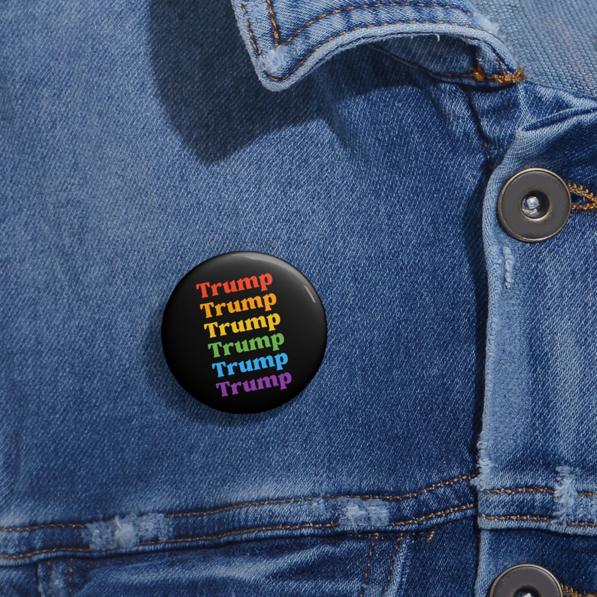 A denim jacket showcases a round metal with Mylar face button, featuring the word Trump repeated in rainbow colors—red, orange, yellow, green, and purple—vertically displayed as an eye-catching nod to LGBTQ Pride.