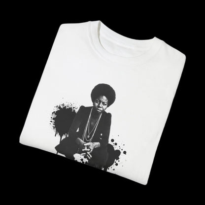 A folded white unisex T-shirt with a vintage look features a black and white print of Nina Simone seated, in front of a  black paint splatter design, set against a black background.