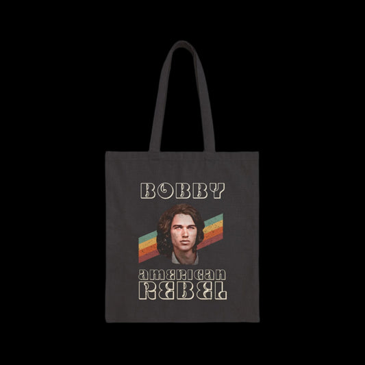 A black cotton canvas tote bag showcases a retro-style image of a long-haired 70's RFK Jr. The text reads BOBBY at the top and AMERICAN REBEL below, set against a striped background in orange, yellow, and green hues reminiscent of the 1970's era.