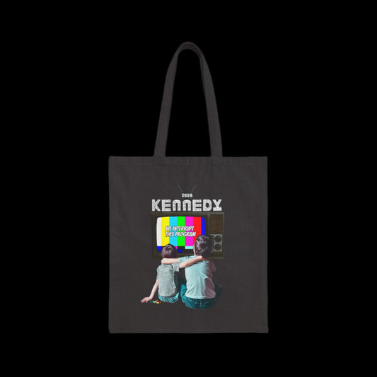 A black cotton canvas tote bag featuring a graphic of two children sitting in front of an old TV displaying a colorful test screen that reads "WE INTERRUPT THIS PROGRAM" Text above reads KENNEDY 2024. The scene has a nostalgic, vintage vibe against a plain black background.