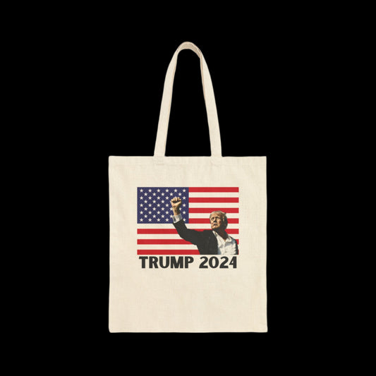 A 100% cotton canvas tote bag featuring an image of Donald Trump  in a suit raising his fist against the backdrop of an American flag, boldly displaying TRUMP 2024. The bags striking design is perfectly highlighted against a sleek black background.