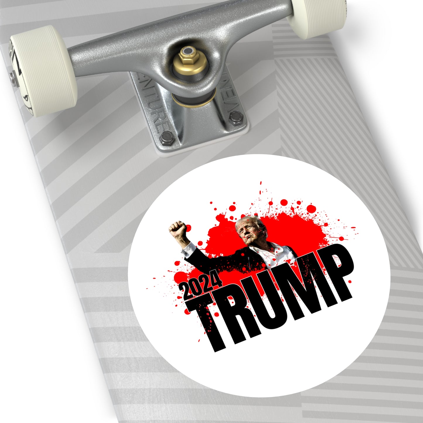 A skateboard with a gray and white striped design features a premium vinyl sticker. The circular Trump sticker shows Donald Trump raising a fist after an assassination attempt with red splatters around and reads 2024 TRUMP.