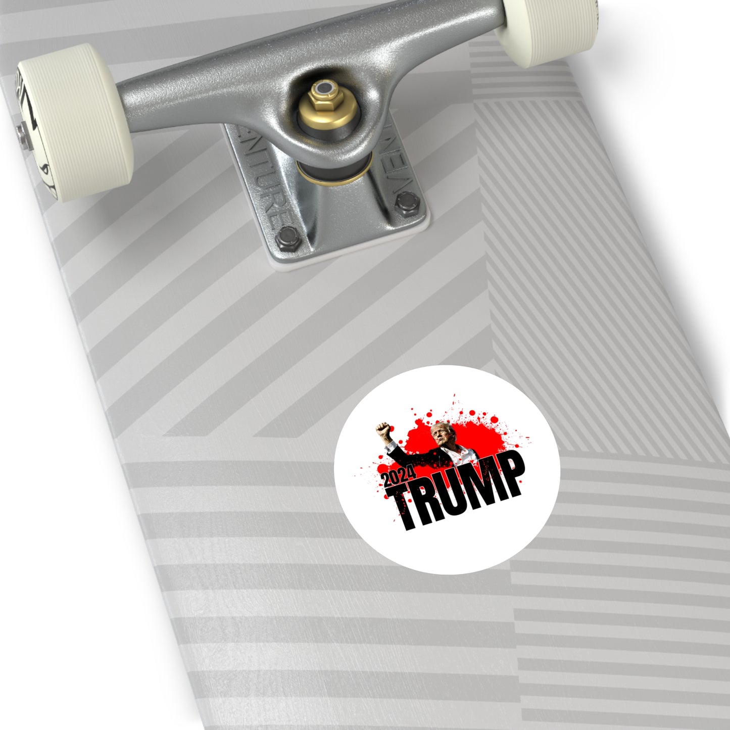 Close-up of a skateboard showcasing a geometric gray pattern. A premium vinyl sticker reads 2024 TRUMP in bold letters, accented by the silhouette of Trump with a raised fist and red splatter effects. The skateboard features white wheels and a silver truck, adding to its striking appeal.