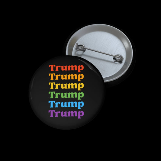 A Rainbow Pin Button features Trump repeated in vibrant hues: red, orange, yellow, green, and purple. Next to it lies an open metal button with a Mylar face, revealing the pin mechanism against a black backdrop.