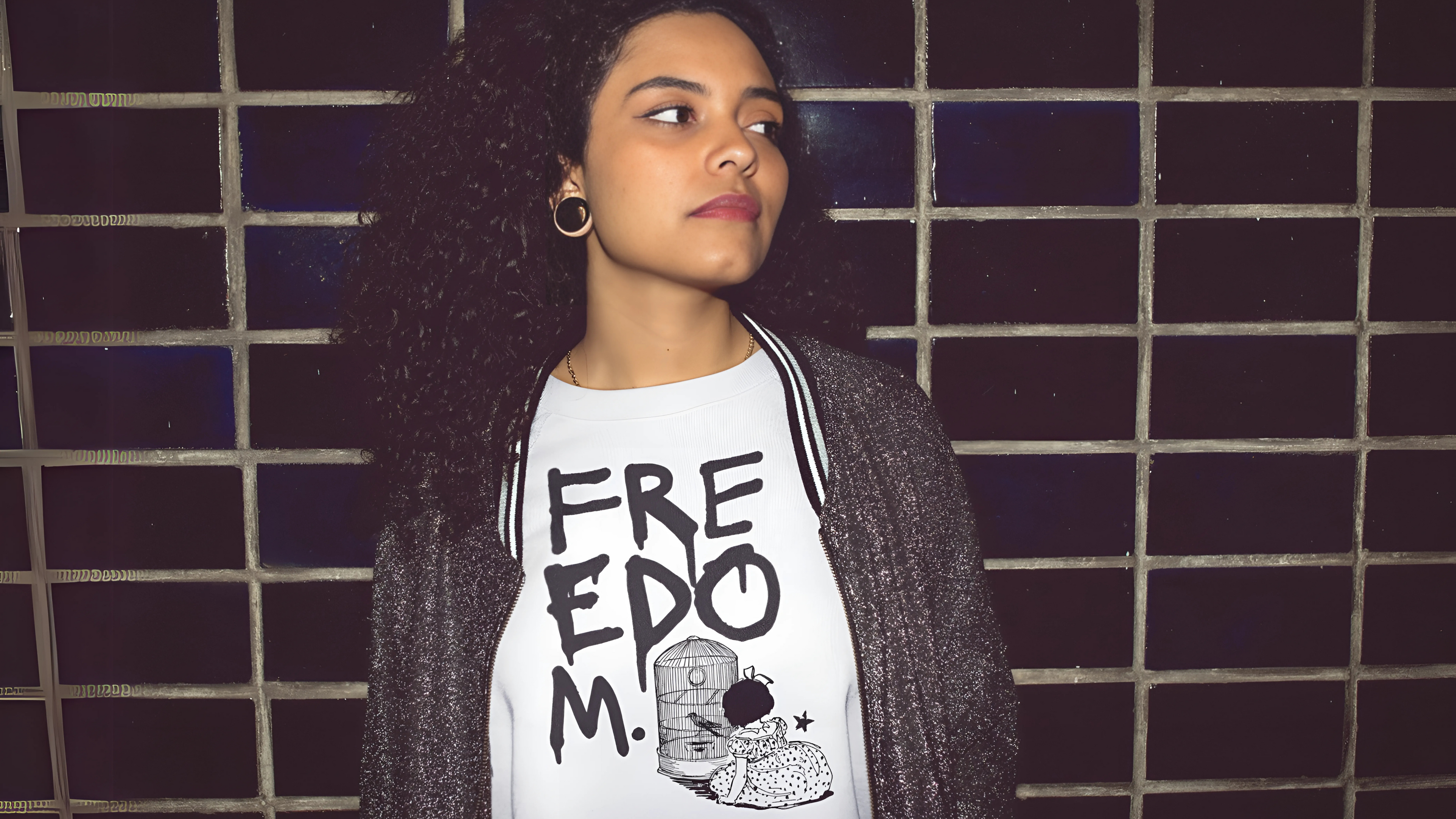 A woman stands against a subway station's blue glazed brick wall. She is looking confidently to the left and wearing a white streetwear tee that says FREEDOM, reminiscent of a brave, strong, rebel woman.