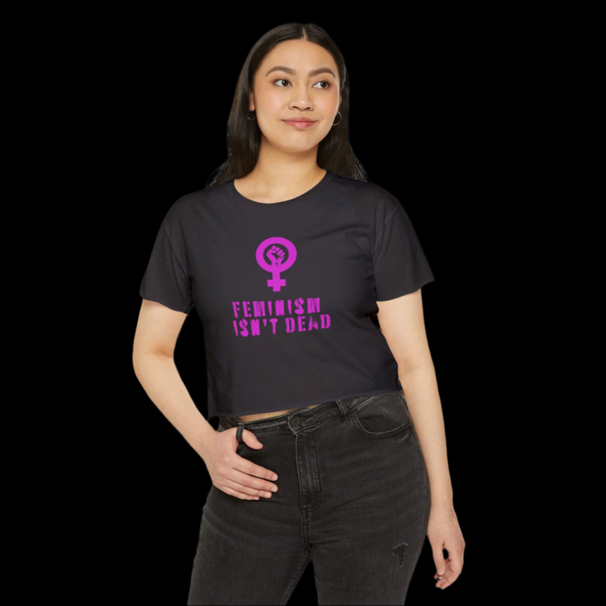 A person with long dark hair, wearing a black crop top emblazoned with pink text and a feminist symbol, stands against a black background. The text reads Feminism Isnt Dead. They have a calm expression and one hand on their hip, embodying the spirit of women's rights.